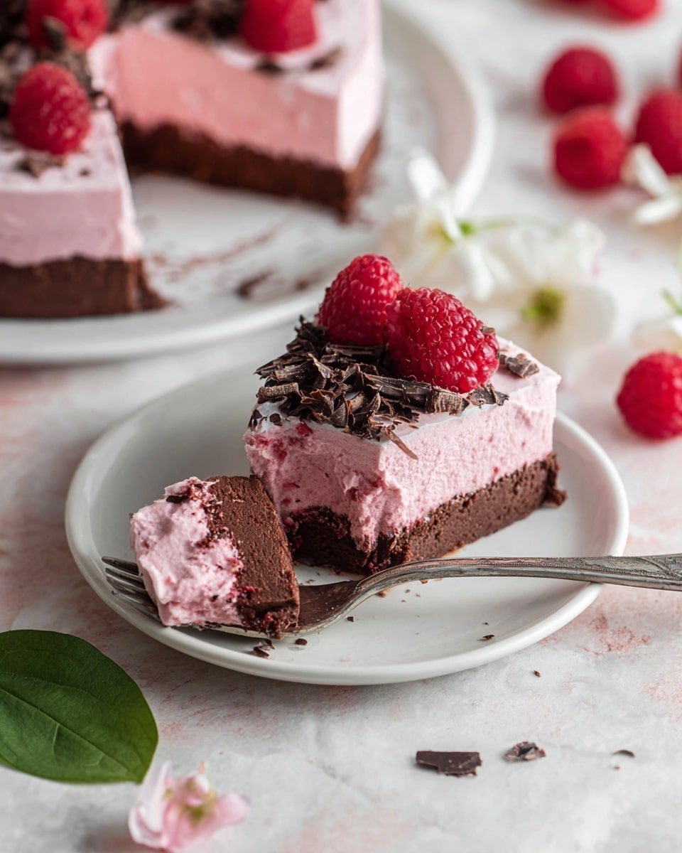 A slice of pink raspberry mousse cake sits on a white plate with a silver fork holding a small bite in front. The cake has two layers: a dark brown thick base layer with a soft texture, and a lighter pink mousse layer on top, smooth and creamy. The mousse layer is decorated with fresh raspberries and thin dark chocolate shavings scattered across the surface. In the background, the rest of the cake with similar pink and brown layers is partly visible on a white plate. White flower petals and bright red raspberries are scattered on the white marbled surface around the plate. A green leaf lies near the plate’s edge. photo taken with an iphone --ar 4:5 --v 7
