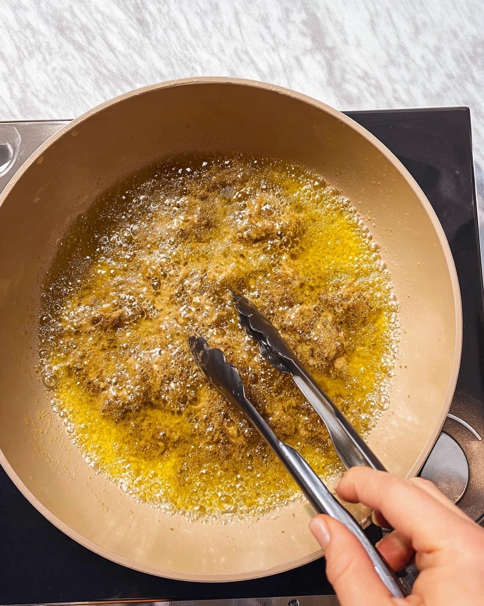 A close-up of small brown pieces frying in hot golden oil inside a light beige pot, with bubbles forming around the food showing the frying process. A woman's hand is holding black and silver metal tongs from the bottom right corner, stirring the pieces in the oil. The pot is placed on a stove, all set on a white marbled surface. Photo taken with an iphone --ar 4:5 --v 7