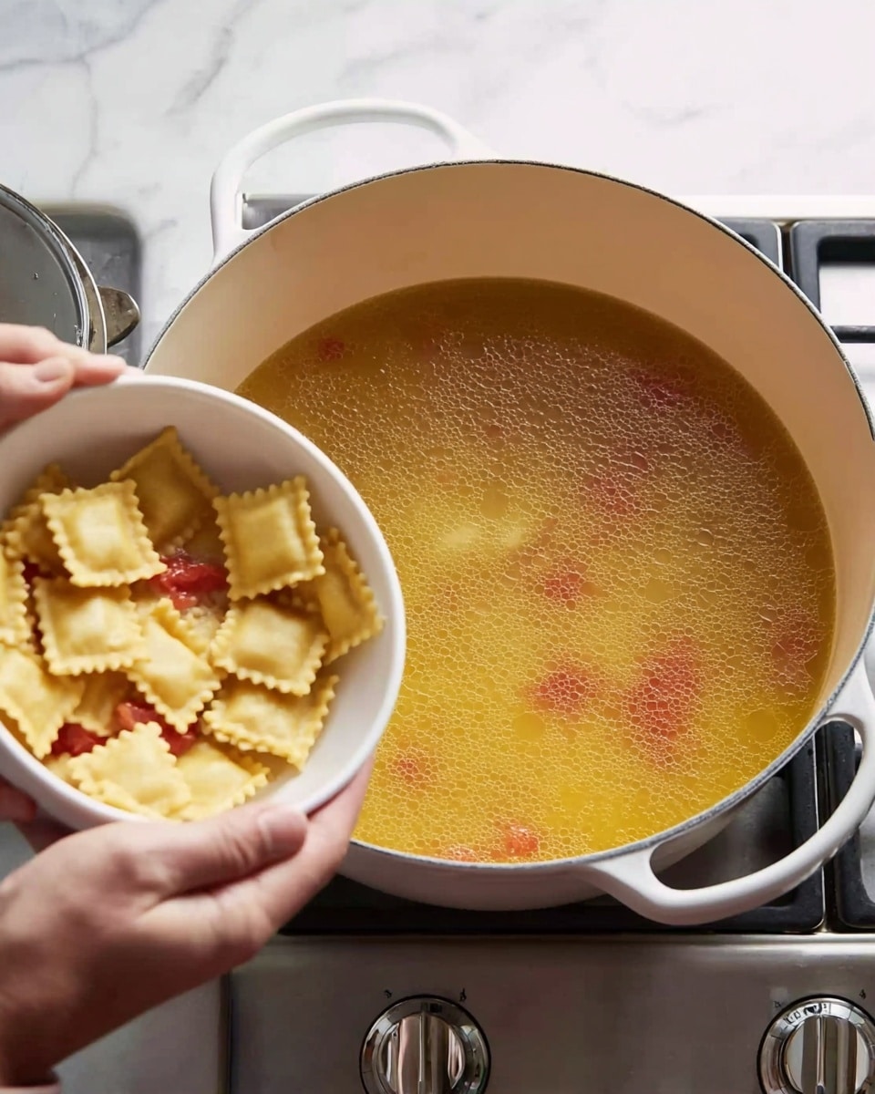 A woman's hand is holding a white bowl above a white pot on a stove, pouring small square ravioli pasta into a light yellow broth with small pieces of red tomato floating in it. The pot is filled almost halfway with the broth, and the scene shows the stove's silver knobs to the right. The background surface is a white marbled texture. Photo taken with an iphone --ar 4:5 --v 7