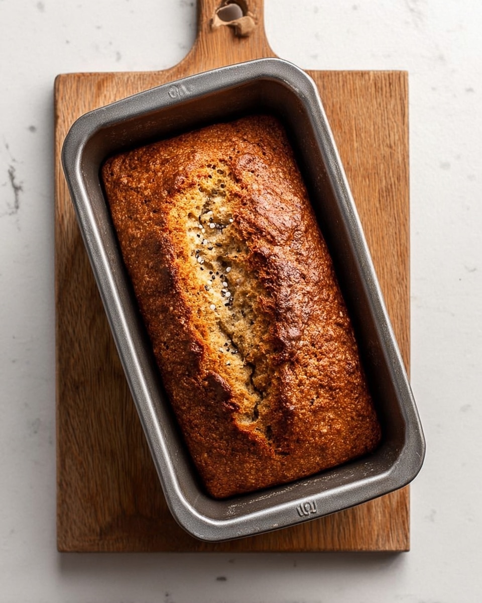 A baked loaf in a gray metal rectangular pan sits on a wooden board with a handle, placed on a white marbled surface. The loaf has a golden brown crust with a rough texture. There is a cracked line in the middle of the loaf showing a denser, lighter inside with small black and white seeds or grains. The surface of the loaf looks dry and firm, with some uneven bumps and a rustic appearance. Photo taken with an iphone --ar 4:5 --v 7