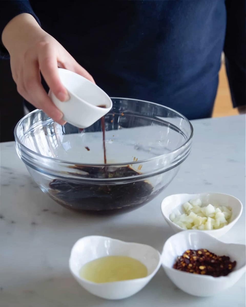 A woman’s hand is pouring a dark brown liquid from a small white bowl into a large clear glass mixing bowl. The dark liquid pools at the bottom of the big bowl. In front of the mixing bowl, there are four small white bowls arranged loosely in a row: the first has a light yellow liquid, the second has chopped white garlic, the third is out of focus, and the fourth holds red pepper flakes. The scene is set on a white marbled surface. The person pouring wears a dark blue long-sleeve shirt. Photo taken with an iphone --ar 4:5 --v 7