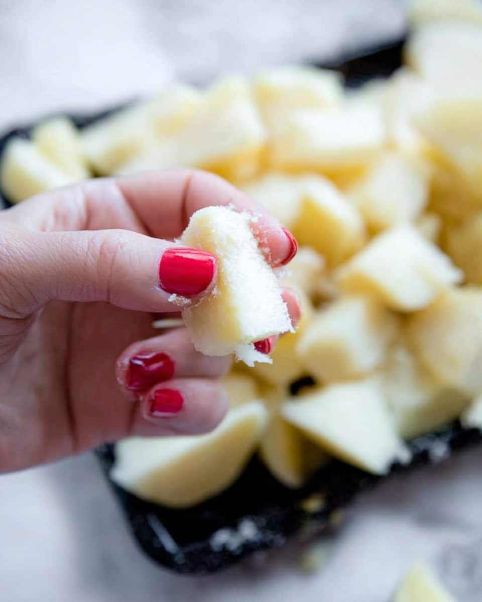 A close-up of a woman's hand with red nail polish holding a torn piece of pale yellow food, likely a soft vegetable or fruit. In the background, there are many similar pieces piled on a dark tray, contrasting with the white marbled surface underneath. The image focuses on the texture and light colors of the torn piece and the fingers holding it. photo taken with an iphone --ar 4:5 --v 7
