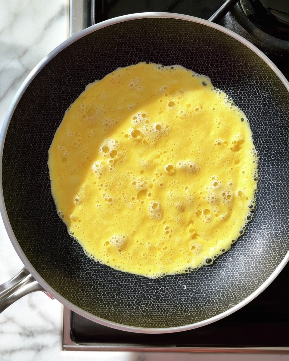 A single layer of yellow beaten egg mixture spread thinly and cooking in a black frying pan with a honeycomb pattern, with small bubbles forming on the surface; the pan has a silver rim and sits on a stovetop, with part of the white marbled countertop visible below. photo taken with an iphone --ar 4:5 --v 7