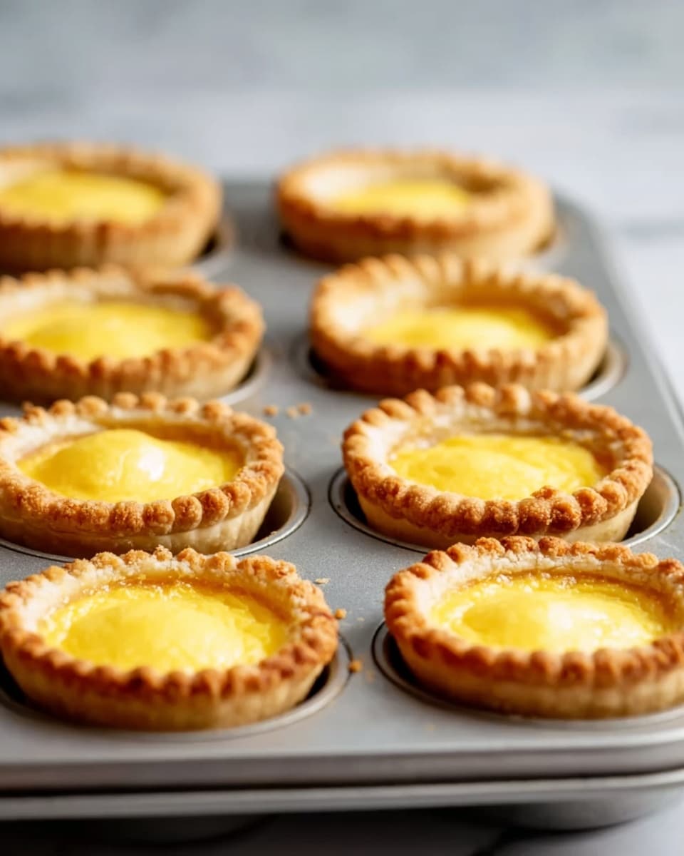 The image shows a tray with nine small pies baked in a metal muffin tin. Each pie has a golden-brown crust with a crimped edge and a smooth, slightly shiny yellow filling in the center. The crust looks flaky and crisp, with a deeper golden color around the edges. The metal muffin tray is placed on a white marbled surface, and the background is softly blurred with neutral colors. The focus is on the front pies, showing clear texture and color contrast between the crust and the filling. photo taken with an iphone --ar 4:5 --v 7