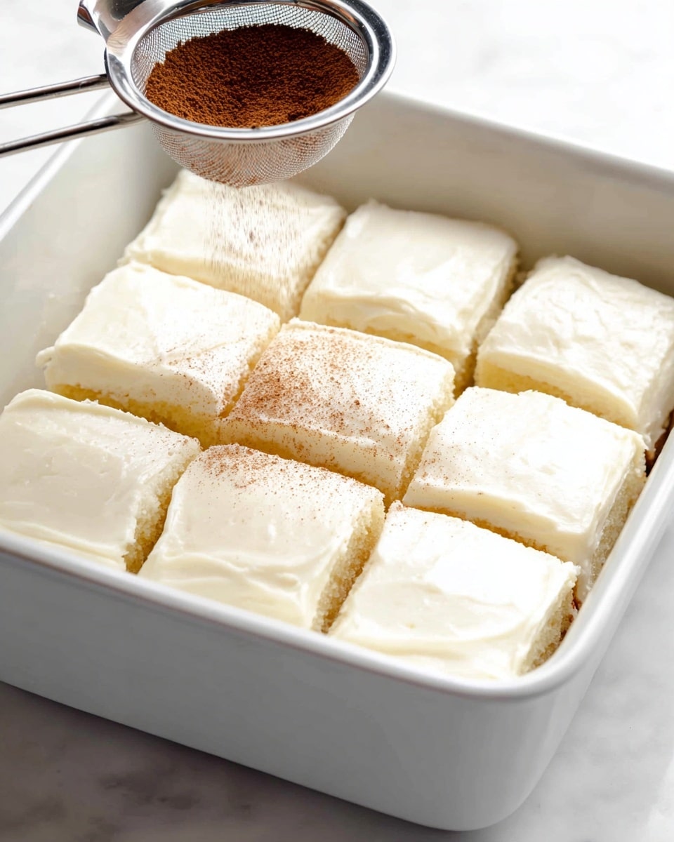 A white ceramic baking dish holds nine square pieces of soft, light yellow cake with smooth, creamy white frosting covering each piece evenly, arranged in three rows and three columns. Above the cake, a silver metal tea strainer is shown sprinkling a fine layer of brown powder, likely cinnamon, over the frosted squares. The background is a white marbled texture. Photo taken with an iphone --ar 4:5 --v 7