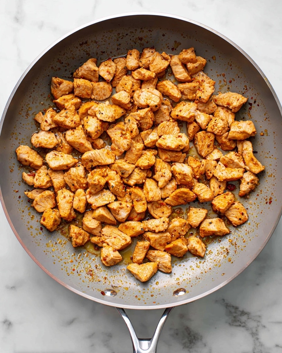 The image shows small, golden-brown pieces of cooked meat spread evenly in a large gray pan with a shiny silver handle. The meat pieces have a slightly rough texture with some darker spots indicating light browning. The pan sits on a white marbled surface, and small splashes of seasoning or sauce speckles are visible on the pan’s inner sides. The scene is well-lit with clear details of the textured meat and pan surface photo taken with an iphone --ar 4:5 --v 7