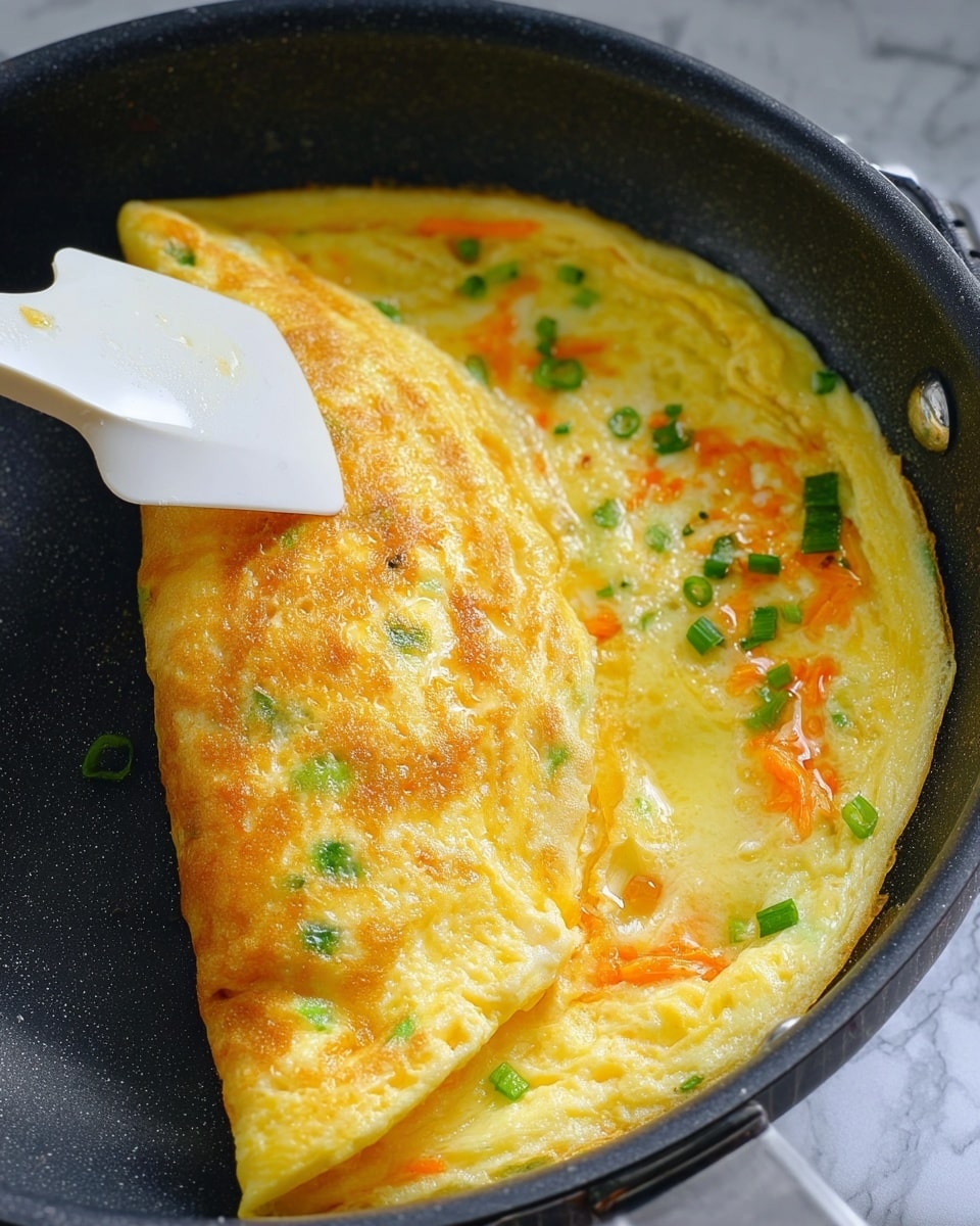 This image shows a close-up of a cooking omelet in a black non-stick pan. The omelet is partially folded in half, with the folded part having a golden brown color with small green bits inside, likely chopped green onions. The unfolded part has a glossy yellow surface with visible bits of grated orange carrot and more chopped green onions spread evenly. A white spatula is lifting the folded part gently. The pan's handle and part of a white marbled surface can be seen in the background. photo taken with an iphone --ar 4:5 --v 7