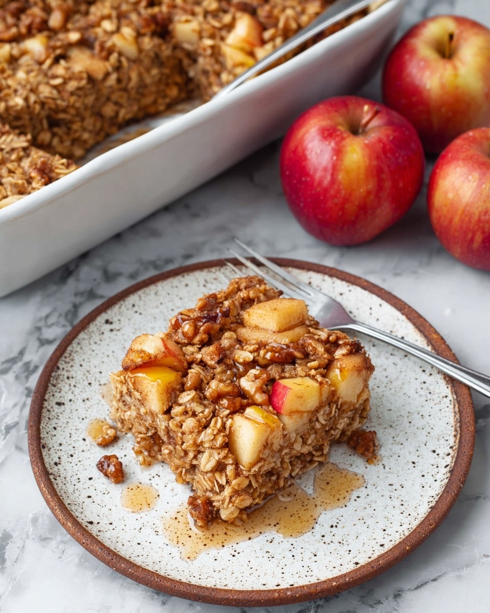 A square piece of baked apple oatmeal sits on a white speckled plate with a brown edge, showing a dense mix of golden brown oats, walnut pieces, and small apple chunks in light red and yellow hues, some syrup drizzled around it. The oatmeal appears soft but textured with a rough oat layer on top. Behind the plate is a white baking dish filled with the same apple oatmeal, revealing a thick, slightly crumbly top layer with visible walnut pieces and diced apples. To the right of the baking dish are two shiny red apples, and a silver fork leans on the plate’s edge. All items rest on a white marbled surface. Photo taken with an iphone --ar 4:5 --v 7