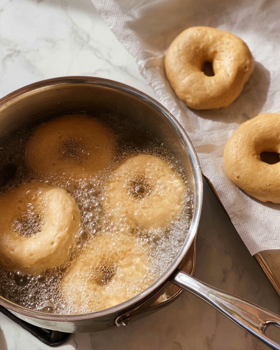 There is a silver pot on a stove with four light brown dough rings boiling in bubbly water, showing a soft, slightly rough texture. To the right, three more dough rings rest on white parchment paper lined on a tray, with a smooth and raw look. The scene is set on a white marbled surface, with the shiny metal pot handle visible on the right side. photo taken with an iphone --ar 4:5 --v 7