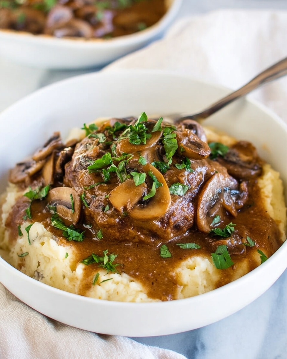 The dish shows a white bowl on a white marbled surface with a serving of mashed potatoes at the base, creamy and slightly chunky in texture with a light off-white color. On top of the potatoes is a thick, round piece of browned meat covered with a rich, dark brown mushroom sauce that includes sliced mushrooms scattered across the top. Fresh green herbs are sprinkled over the meat and sauce, adding a vibrant pop of color. Behind the bowl, a fork rests inside, hinting at readiness to eat. Photo taken with an iphone --ar 4:5 --v 7
