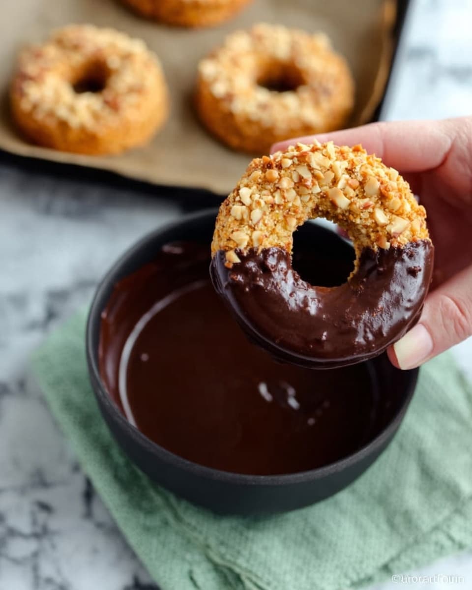 The image shows a close-up of a woman's hand holding a round donut-shaped cookie that is half dipped in smooth dark chocolate. The cookie is light brown with a nutty texture on top and a hole in the center. Below, there is a black bowl filled with melted dark chocolate sitting on a light green cloth. To the left, a baking tray with parchment paper holds two more cookies of the same type, golden brown with a crunchy nut topping. The background surface is a white marbled texture. photo taken with an iphone --ar 4:5 --v 7