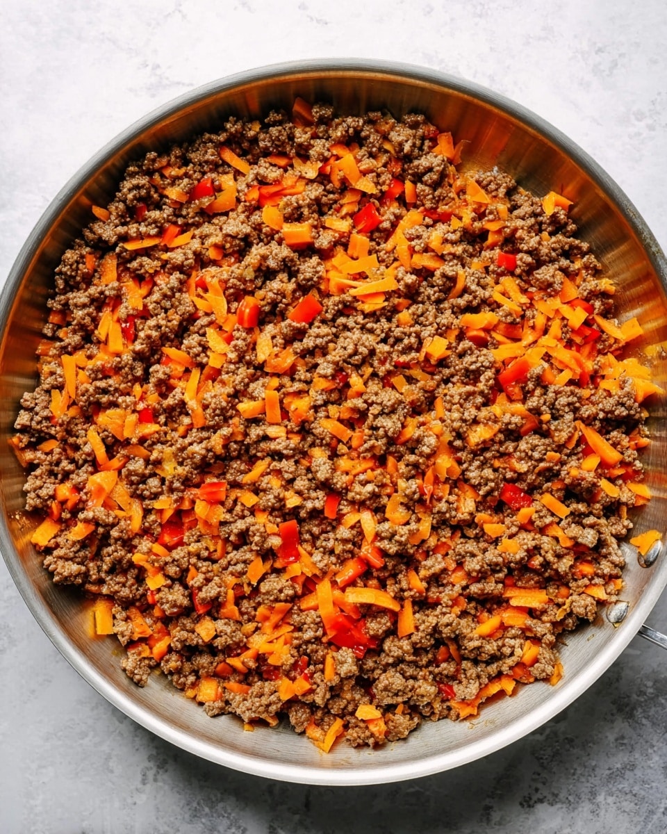 The image shows a silver metal pan filled with cooked ground beef mixed with small chopped orange carrots and red bell peppers, evenly spread across the pan surface. The beef is crumbly and brown, creating a textured base layer, with the bright orange and red vegetable pieces mixed throughout adding pops of color. The pan rests on a white marbled surface, giving a clean and simple background. photo taken with an iphone --ar 4:5 --v 7