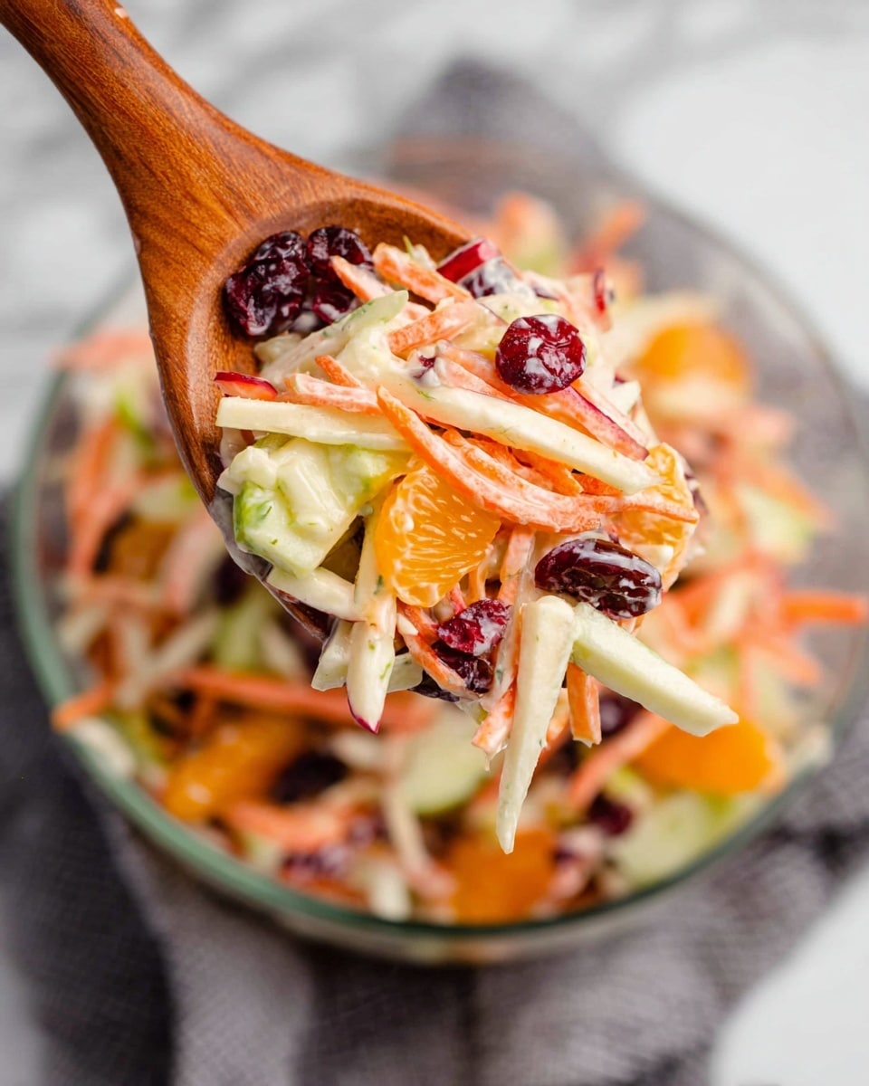 A wooden spoon holds a colorful salad with thin orange carrot sticks, white apple slices with red edges, bright orange mandarin segments, dark red cranberries, and light green cucumber sticks all mixed together with a creamy dressing. The spoon is hovering over a clear glass bowl filled with the same salad on a white marbled surface. A gray cloth is partially visible under the bowl. The focus is on the spoon with the background softly blurred. photo taken with an iphone --ar 4:5 --v 7