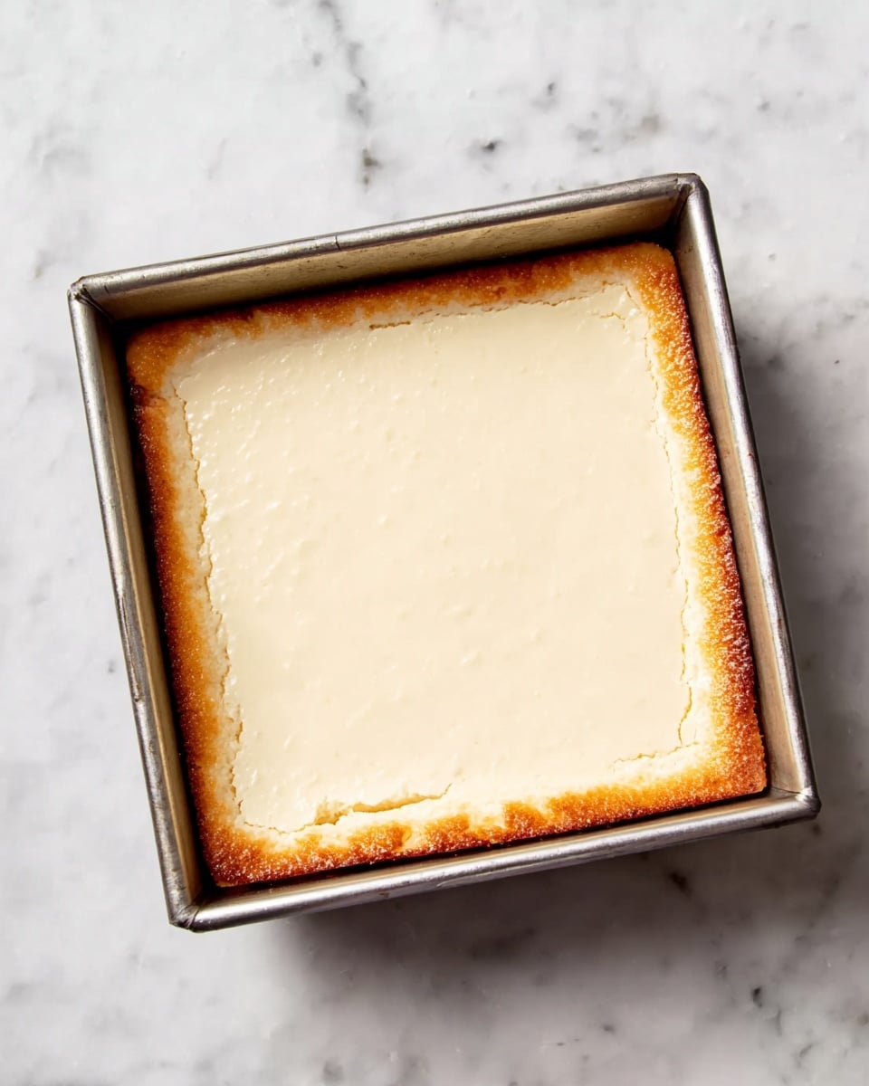 A square metal baking pan sits on a white marbled surface, holding a baked dessert with two visible layers: the bottom layer is golden brown with a slightly crisp edge, and the top layer is a smooth, creamy off-white layer that covers the entire surface evenly. The edges of the top layer show a bit of the golden brown beneath. Photo taken with an iphone --ar 4:5 --v 7