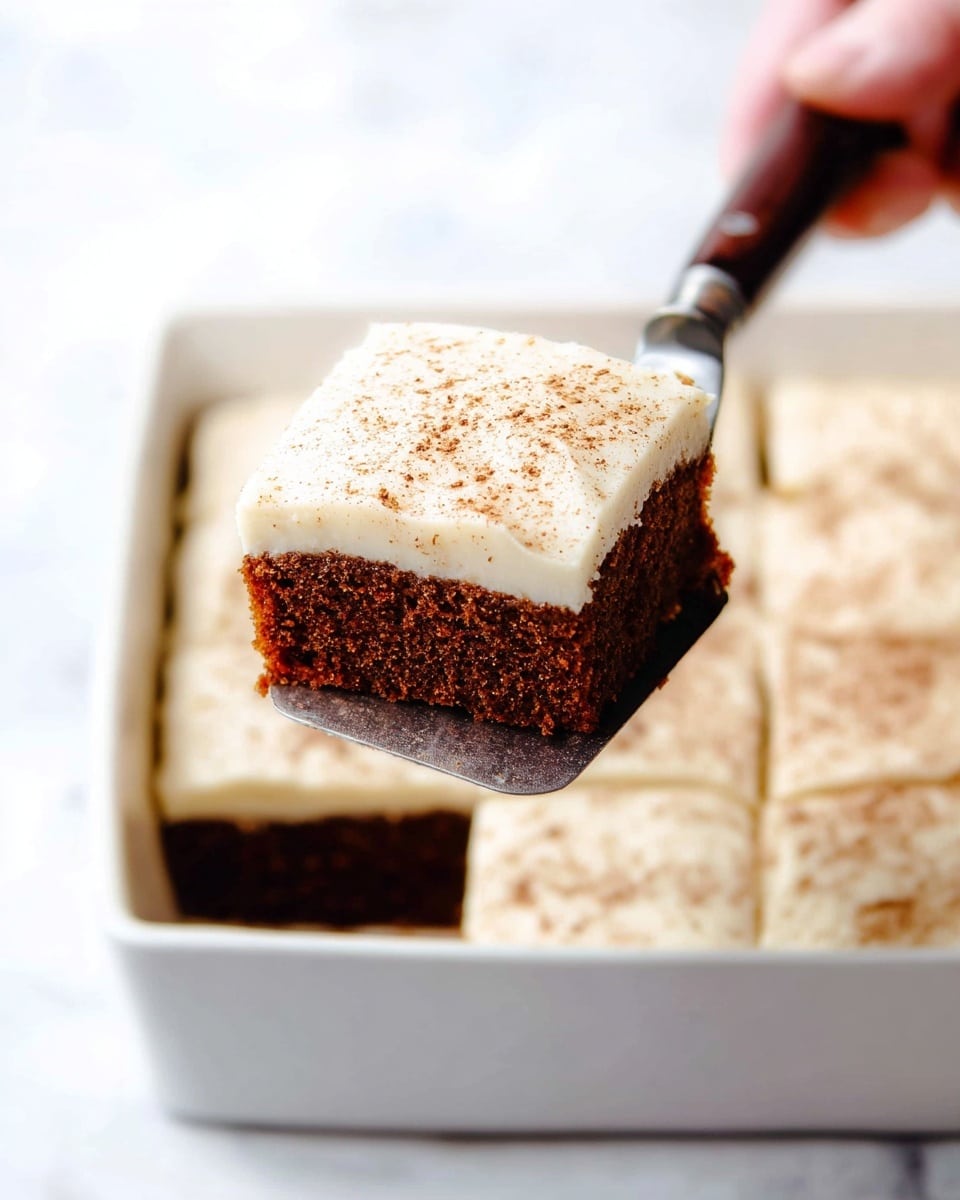 A square piece of dark brown cake with a soft texture is lifted by a metal spatula with a dark wooden handle held by a woman's hand. The cake has one thick layer of smooth, creamy white frosting topped with a light sprinkle of brown powder. The cake piece is shown above a white rectangular dish filled with similar cake squares. The background is a white marbled surface. photo taken with an iphone --ar 4:5 --v 7