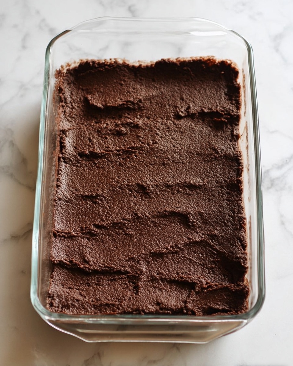 A clear glass rectangular baking dish filled with one thick layer of dark brown chocolate dough spread evenly across the entire surface. The dough has a slightly rough texture with some small uneven spots and gentle ridges. The dish is placed on a white marbled surface, with soft natural light highlighting the texture of the chocolate dough. Photo taken with an iphone --ar 4:5 --v 7