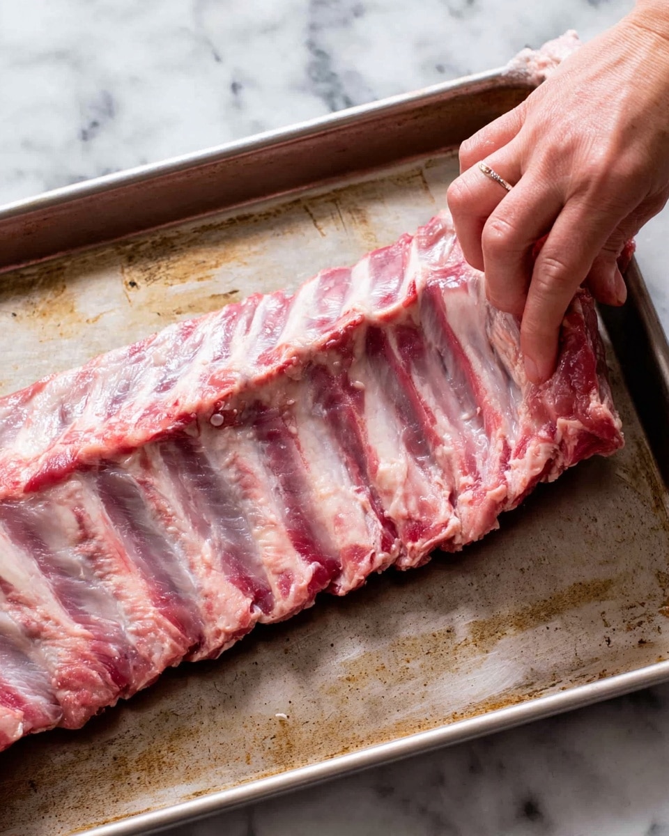 A close-up image shows a raw rack of ribs placed on a metal tray, with the ribs spread out horizontally. The ribs have a pinkish-red color with visible bones running along the length. A woman's hand is lifting a thin, translucent membrane from the top right part of the ribs, pulling it away from the meat. The metal tray beneath the ribs has a worn, slightly stained look. The background is a white marbled texture. photo taken with an iphone --ar 4:5 --v 7