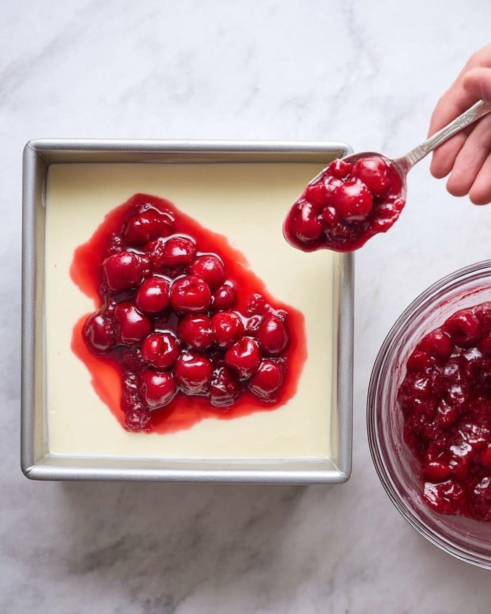 The image shows a square metal pan filled with a smooth, creamy white layer that covers the whole base. A woman's hand holds a spoonful of bright red cherry topping with whole cherries and syrup, hovering over the top right corner of the creamy layer, about to spread it. Next to the pan on the right side, there is a clear glass bowl full of the same red cherry topping. The background is a white marbled surface. Photo taken with an iphone --ar 4:5 --v 7