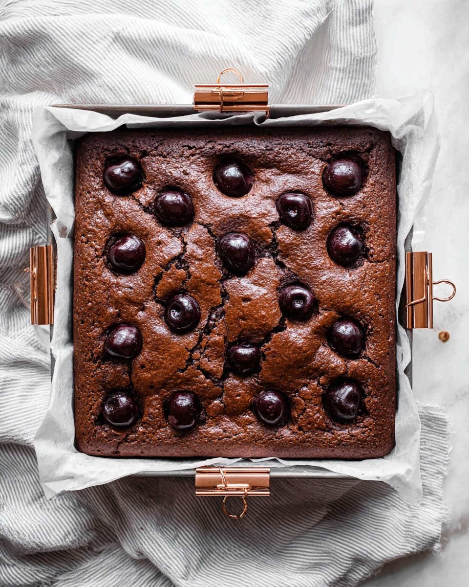 A square chocolate cake with a cracked surface, baked in a metal tray lined with white parchment paper, topped with whole and halved dark cherries pressed into the top. The cake looks dense and moist with a rich dark brown color. The tray is held in place with four rose gold clips on each corner, and it is placed on a crumpled light striped cloth over a white marbled surface. photo taken with an iphone --ar 4:5 --v 7