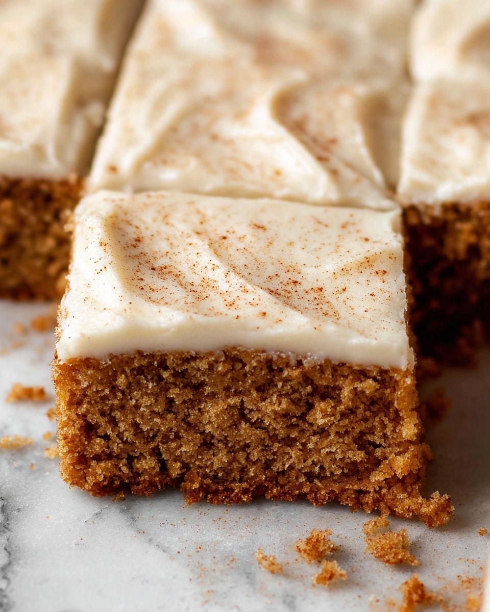 A close-up of a square piece of soft, brown cake with a smooth, creamy white frosting layer on top, sprinkled lightly with cinnamon or nutmeg powder. The cake looks moist and crumbly, revealing its texture where a piece has been separated from the rest. The cake rests directly on a white marbled surface with some crumbs scattered around. The frosting layer is thick, evenly spread, and slightly textured with gentle swirls. Photo taken with an iphone --ar 4:5 --v 7