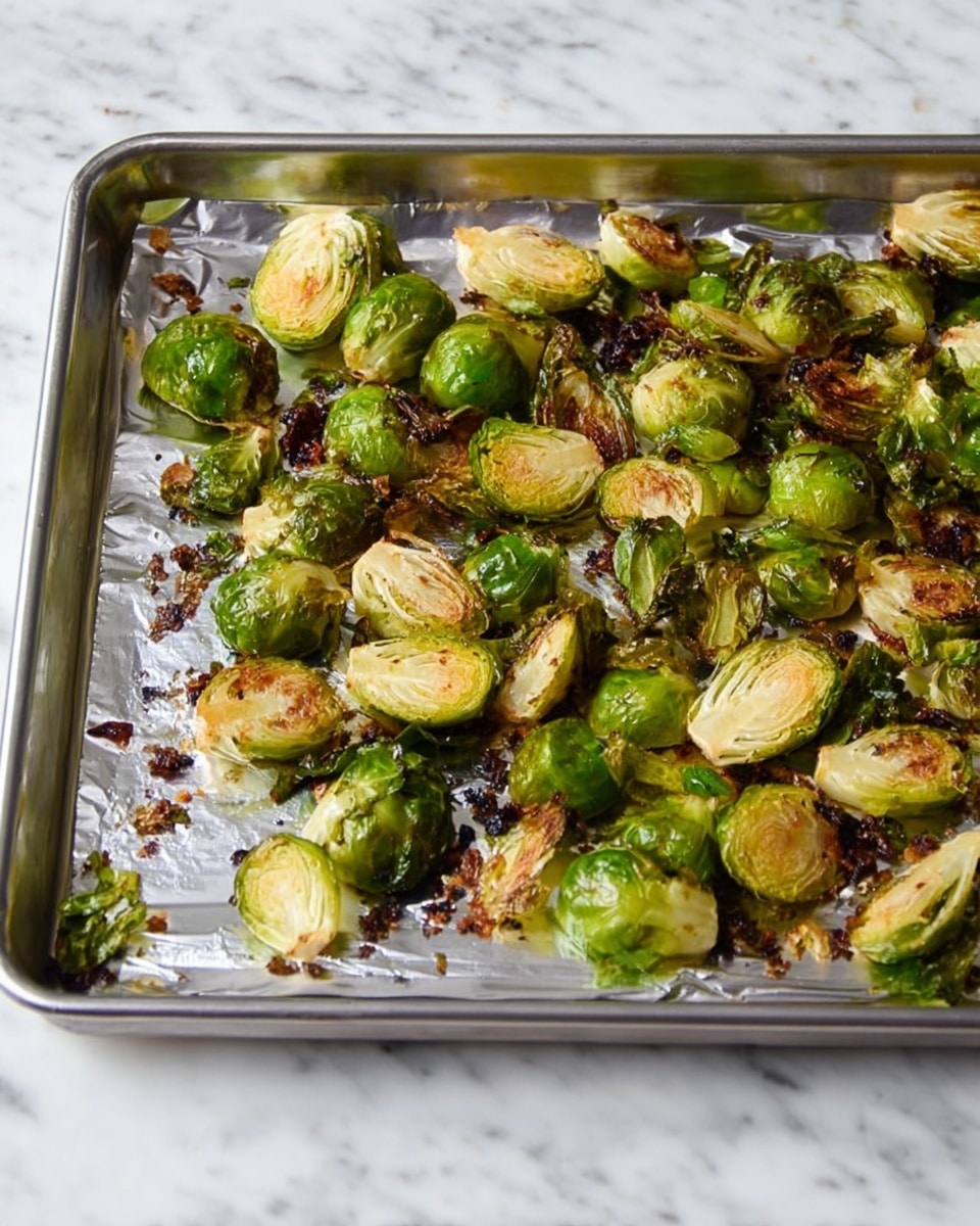 The image shows a baking tray lined with silver foil, filled with roasted Brussels sprouts. The Brussels sprouts are cut into halves, with some halves showing a golden brown, slightly crispy texture while others remain a bright green color. The tray has small bits of browned and slightly charred pieces scattered around, adding to the roasted look. The tray is placed on a clean white marbled surface. Photo taken with an iphone --ar 4:5 --v 7