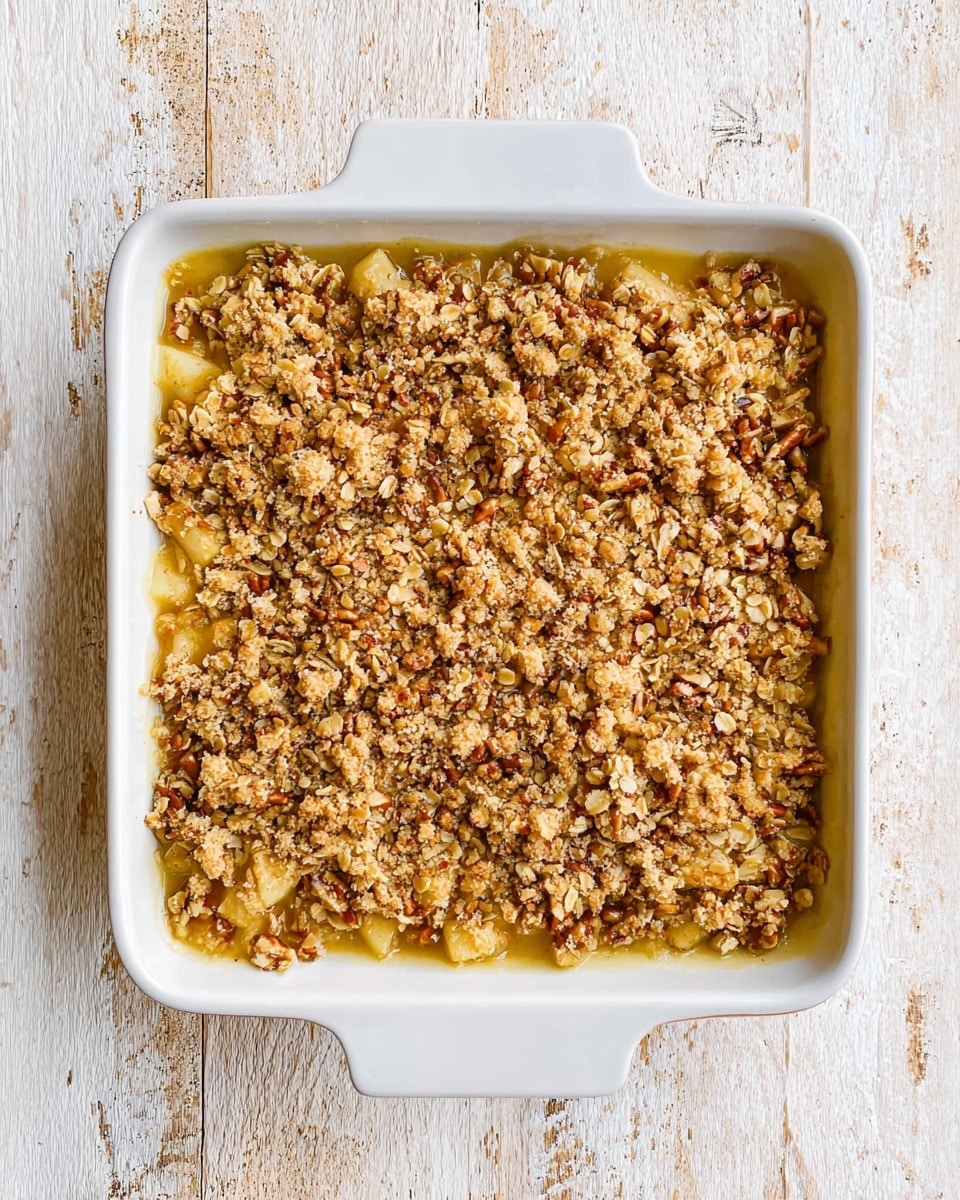 A square white baking dish filled with two visible layers: the bottom layer is soft and light yellow, likely cooked fruit or a sauce, while the top layer is a chunky, golden brown oat crumb topping mixed with pecans and oats, giving it a rough texture. The whole dish sits on a white marbled surface with some rustic wood patterns visible beneath, and the shot is taken from above, showing the full top view of the dessert. photo taken with an iphone --ar 4:5 --v 7