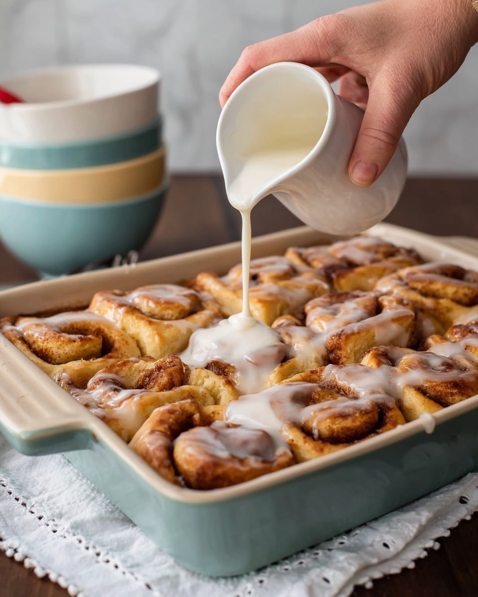 A close-up view of a rectangular baking pan filled with many golden-brown cinnamon rolls arranged tightly side by side. The cinnamon rolls have a slightly crispy texture with visible spiral layers and cinnamon swirls. A woman's hand is pouring thick white icing from a small white pitcher over the cinnamon rolls, creating a shiny, smooth layer of glaze spreading over the top. The pan sits on a white cloth with simple small cross stitches, placed on a dark wooden surface with a soft white marbled background. In the background, blurred, there are stacked pastel bowls in white and light blue with cream-colored rims. Photo taken with an iphone --ar 4:5 --v 7