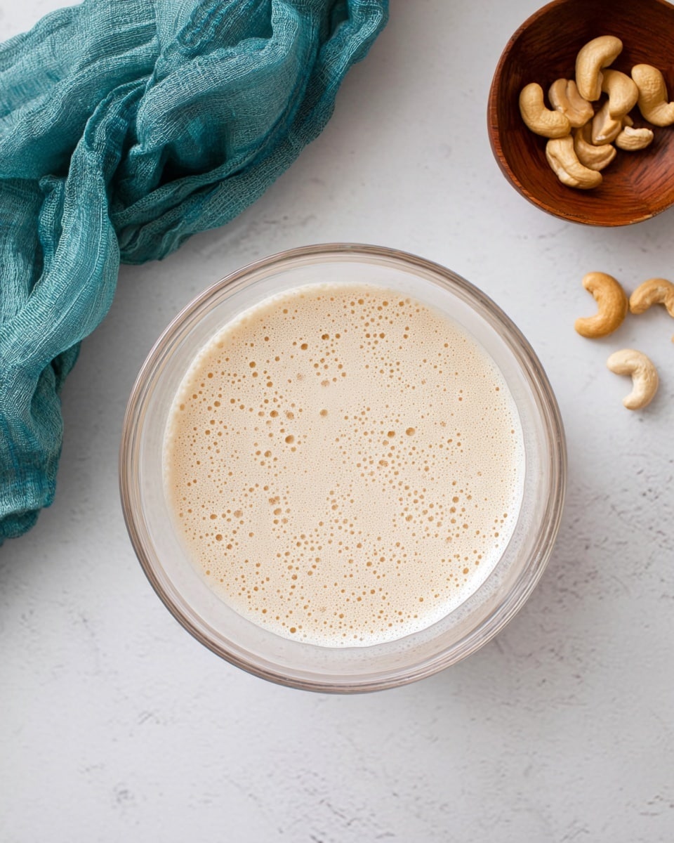 The image shows a clear glass bowl filled with a light beige, frothy liquid that has small bubbles spread evenly across the surface, sitting on a white marbled surface. To the upper right of the bowl, there is a small wooden bowl containing a few light brown cashew nuts. On the left side, there is a soft, crumpled teal fabric piece resting on the white marbled texture. The scene has a clean, minimal look with gentle natural lighting. photo taken with an iphone --ar 4:5 --v 7