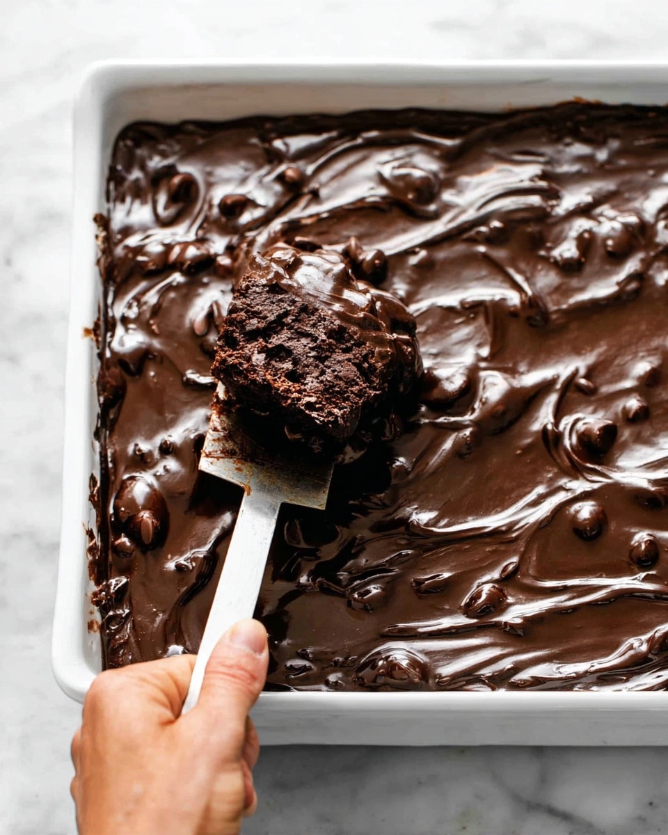 A close-up of a white rectangular dish filled with a rich, thick dark chocolate dessert. The top layer is glossy and textured with uneven swirls and chocolate pieces giving it a bumpy surface. A woman's hand holds a spatula lifting a large, dense chunk from the corner, showing a moist, dark inside that contrasts slightly with the shiny top. The dish rests on a white marbled surface. photo taken with an iphone --ar 4:5 --v 7