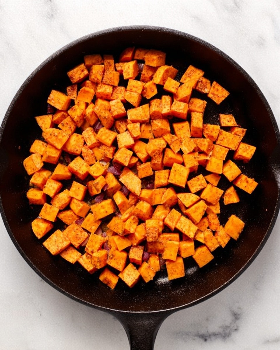 A cast iron pan filled with small cubes of bright orange sweet potatoes, evenly spread across the surface with a few lightly browned edges showing some cooking. The pan is on a white marbled surface, and the sweet potatoes have a slightly rough texture with a warm, almost golden seasoning dusted over them. The pan handle points towards the bottom middle of the image. photo taken with an iphone --ar 4:5 --v 7