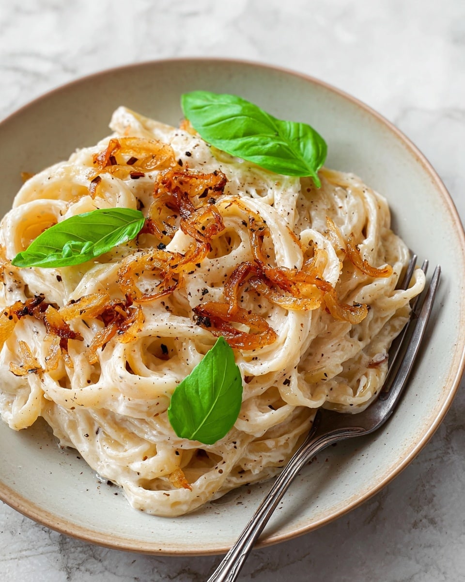 A white plate holds a nest of creamy white pasta layered thickly with a smooth texture, topped with caramelized golden-brown onions scattered across and mixed in. The pasta is sprinkled with coarse black pepper and garnished with two fresh bright green basil leaves, one larger on the left side and a small one in the center. A silver fork rests on the plate’s edge partially under the pasta. The background is a white marbled surface. photo taken with an iphone --ar 4:5 --v 7