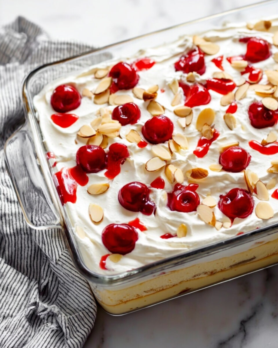 A clear glass rectangular dish holds a dessert with three visible layers: the bottom layer is light and fluffy cream, the middle layer is soft and pale yellow cake, and the top layer is white whipped cream spread smoothly with slight swirls. Red cherries, some with a little syrup, are scattered evenly across the top cream layer along with thin, light brown almond slices. The dish rests on a white marbled surface with a striped cloth beside it. photo taken with an iphone --ar 4:5 --v 7