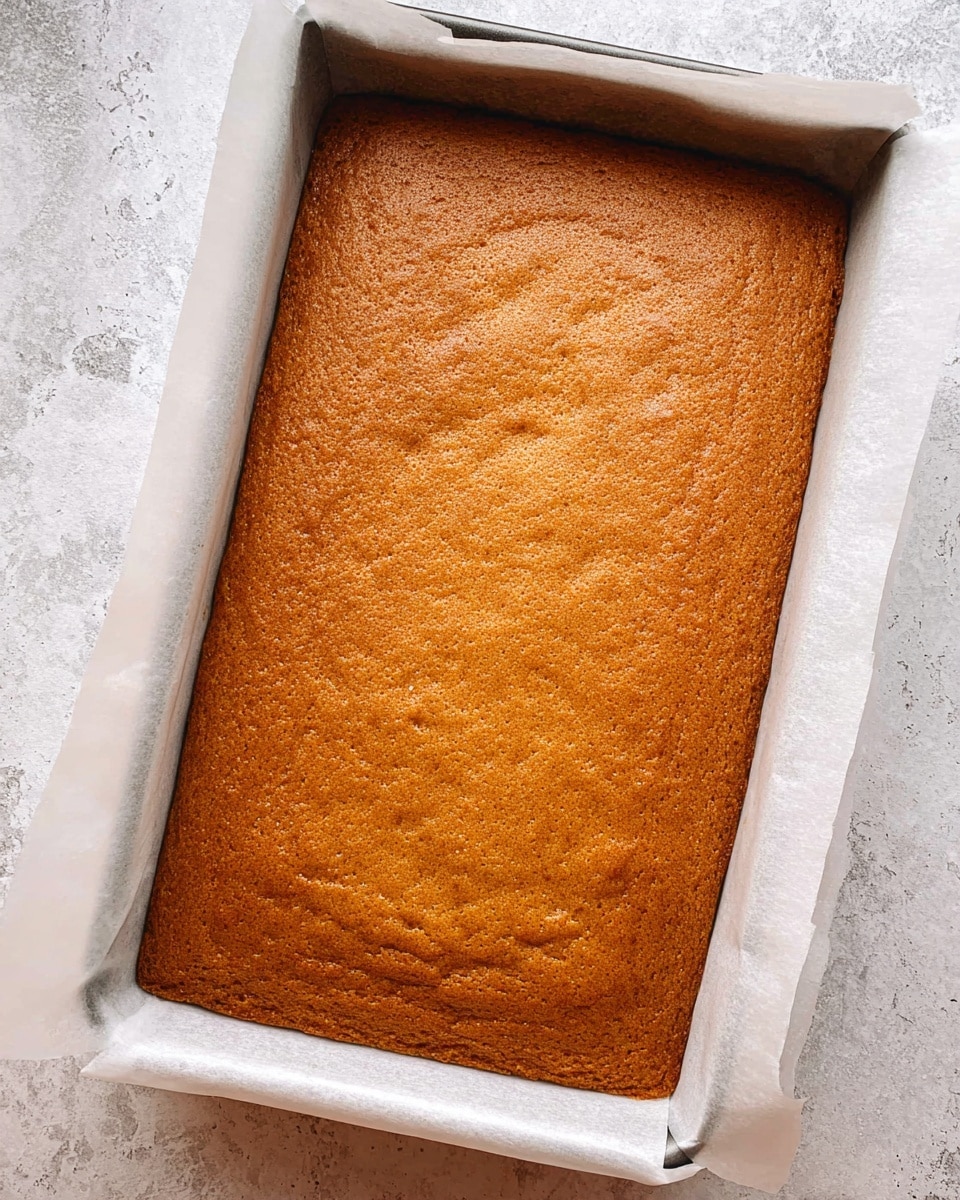A large, rectangular single-layer golden-brown cake with a slightly rough texture rests inside a baking tray lined with white parchment paper that lifts slightly at the edges. The surface of the cake is even with a few small dimples and a matte finish. The baking tray sits on a white marbled textured surface. Photo taken with an iphone --ar 4:5 --v 7