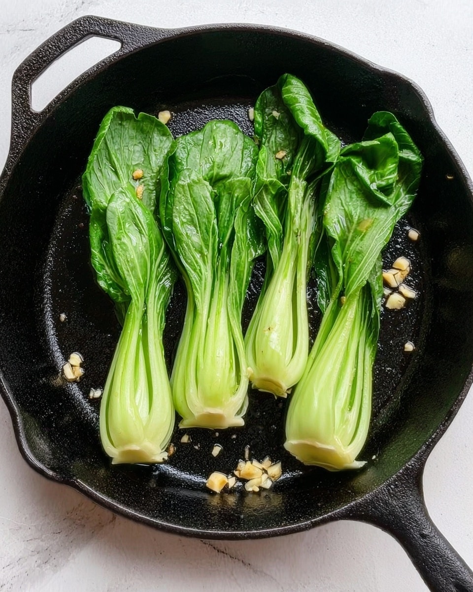 The image shows a black cast iron skillet on a white marbled surface. Inside the skillet, there are four bright green bok choy pieces placed side by side, with their white stems at the bottom and leafy green tops pointing upward. Small pieces of chopped garlic are scattered around the bok choy on the skillet, adding texture and detail. The overall look is fresh and simple, focusing on the vibrant greens against the dark pan. Photo taken with an iphone --ar 4:5 --v 7