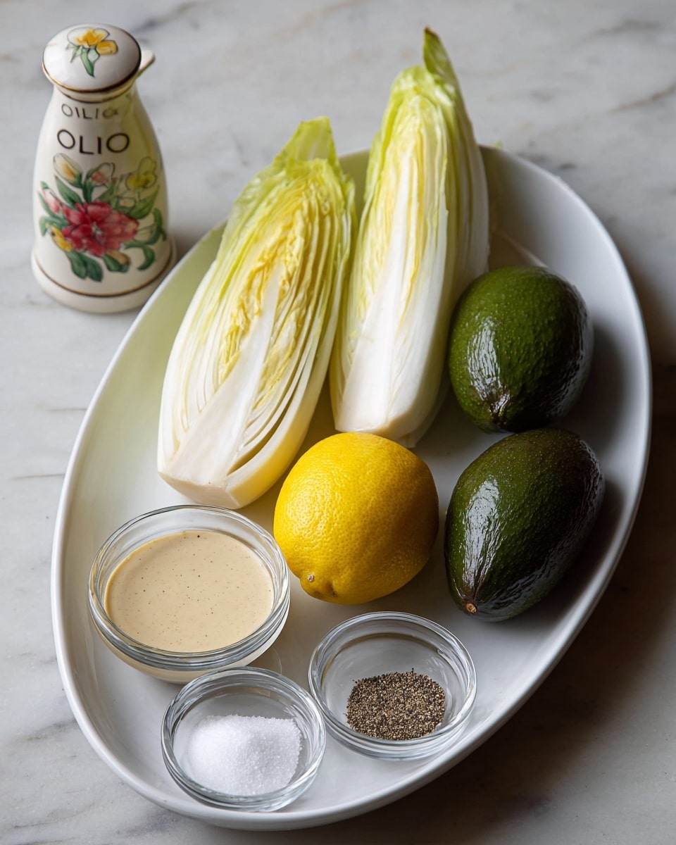A white oval plate sits on a white marbled surface holding several fresh ingredients: two pale yellow and white endives on the left, a bright yellow lemon in the center front, two dark green avocados on the right, a small clear glass bowl of light beige sauce on the left front, and two smaller clear glass bowls with black pepper and white salt placed front center. In the background, a small white ceramic container with floral designs and the word