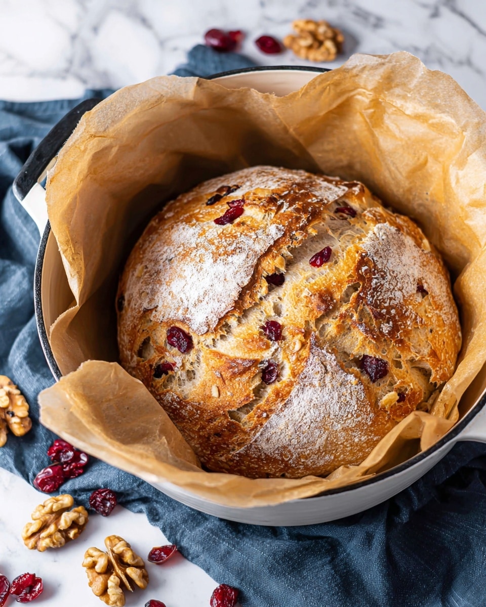 The image shows a round loaf of bread with a golden-brown crust resting inside a white pot lined with light brown parchment paper. The bread has a cracked surface dusted with white flour, revealing bits of red cranberries and light brown walnuts embedded inside. Around the pot on the white marbled surface, there are scattered whole walnuts and dried cranberries. A dark blue cloth is partially visible beneath the pot. photo taken with an iphone --ar 4:5 --v 7
