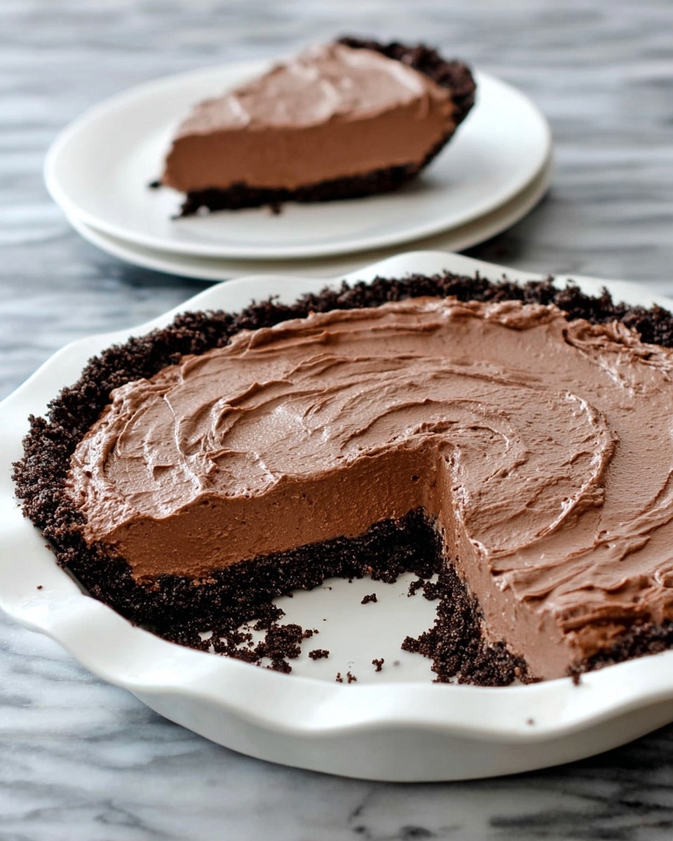 A large chocolate pie is shown in a white plate with a wavy edge, with one slice missing and some crumbs scattered near the empty space. The pie has two clear layers: a dark crumbly chocolate crust at the bottom and a thick, creamy chocolate filling on top with a smooth, slightly swirled texture. In the background, a slice of the same pie is placed on a smaller white plate. The whole setup sits on a white marbled surface. photo taken with an iphone --ar 4:5 --v 7