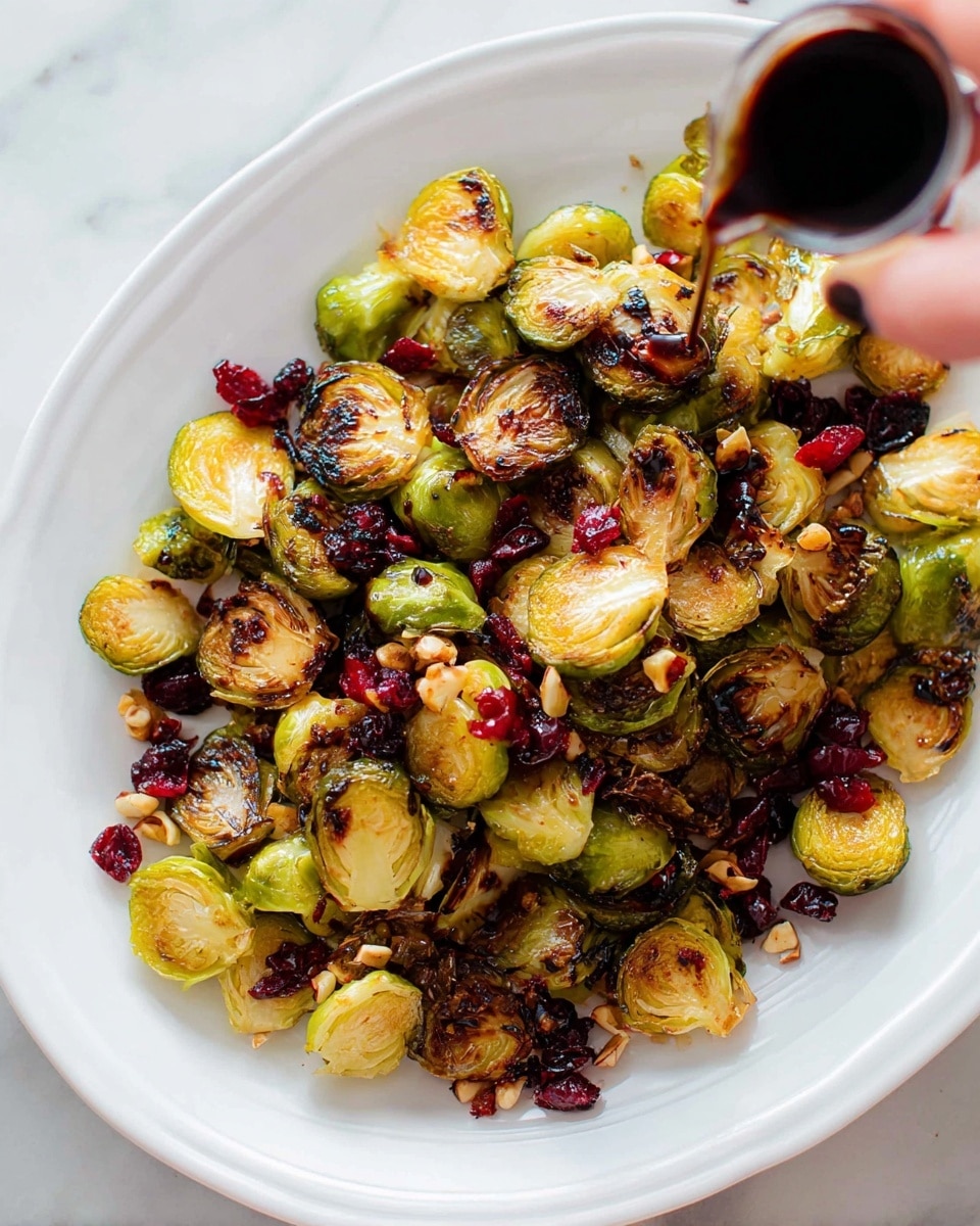 The dish shows a white plate filled with roasted Brussels sprouts that are golden brown and slightly charred, cut in half showing and radiating their green and light yellow layers. Scattered over and mixed between the Brussels sprouts are small pieces of red dried cranberries and some brown chopped nuts for texture. A dark balsamic glaze is being poured over the top from the side of the image, adding a shiny layer to the dish. The plate sits on a white marbled surface and a woman's hand holding the small container of glaze is visible in the corner. photo taken with an iphone --ar 4:5 --v 7