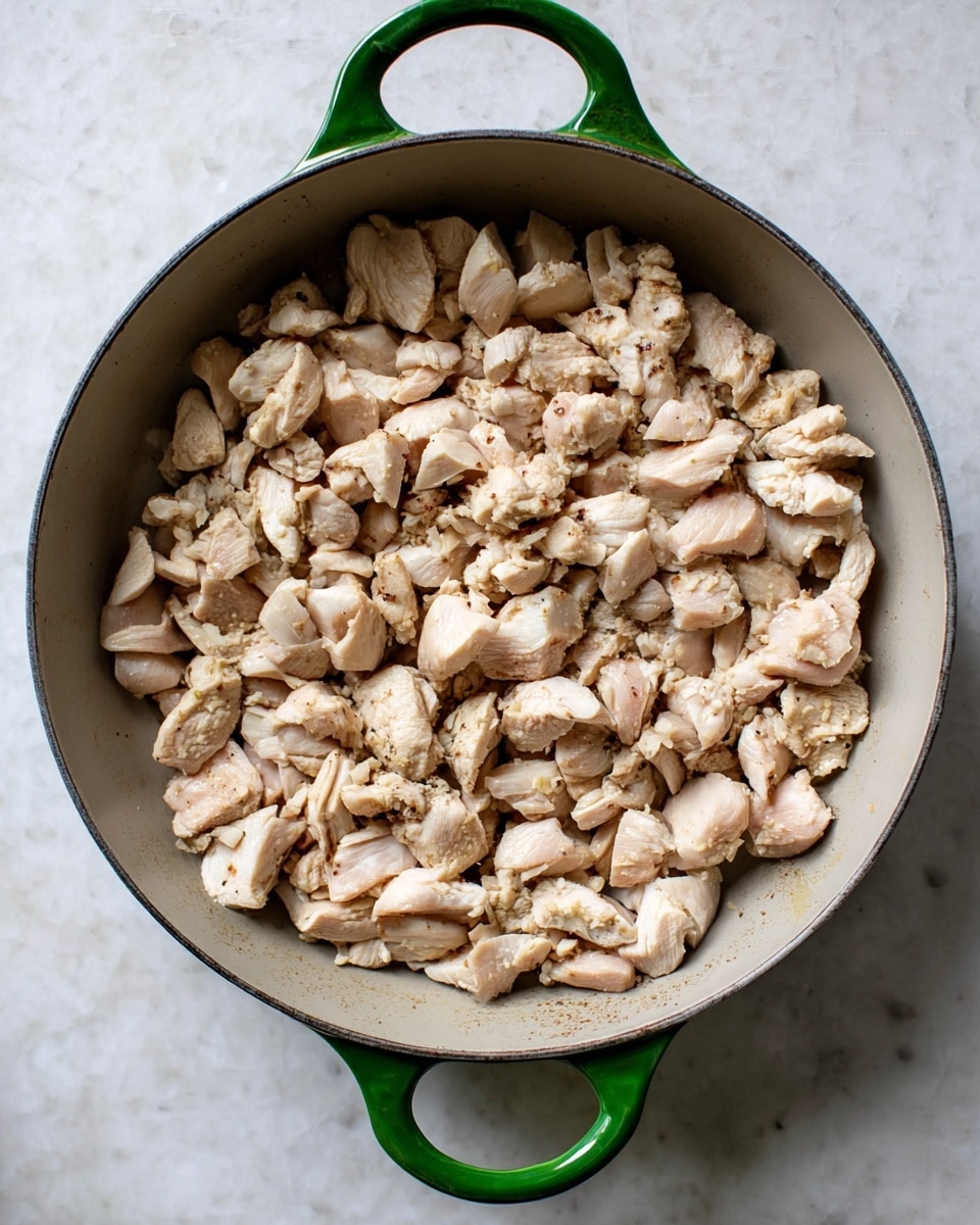 A round pan with a green handle holds many small, light beige cooked chicken pieces spread evenly inside. The chicken pieces have a slightly moist texture with some hints of light brown spots, showing they are lightly cooked. The pan is placed on a white marbled surface, giving a clean and simple background. Photo taken with an iphone --ar 4:5 --v 7