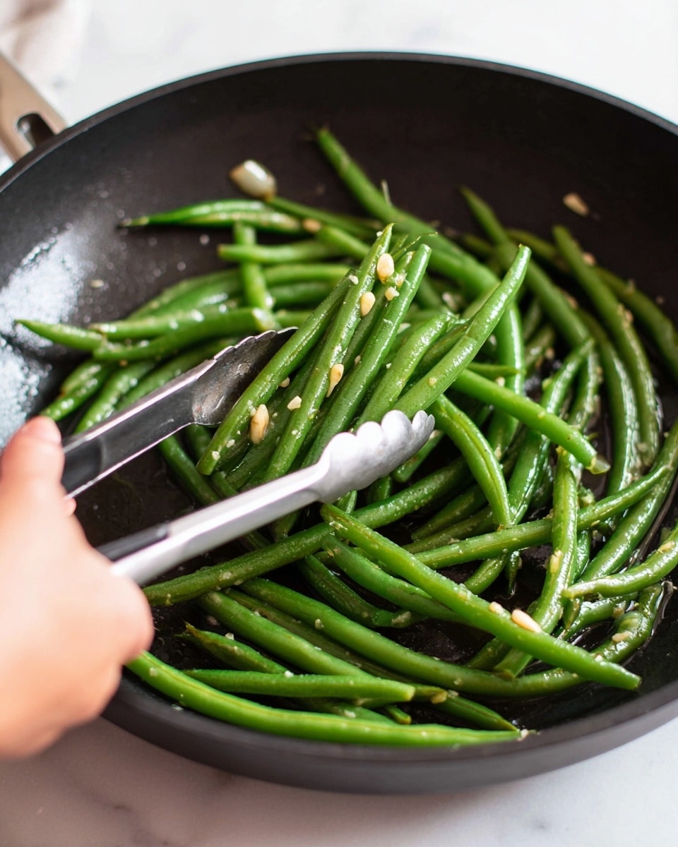 A close-up view of a black frying pan filled with shiny cooked green beans, each bean long and slender with a slight oil glaze, showing small pieces of garlic scattered throughout. A woman's hand is holding silver tongs with black tips, lifting a small bunch of green beans from the pan. The frying pan rests on a white marbled surface, visible in the background. photo taken with an iphone --ar 4:5 --v 7