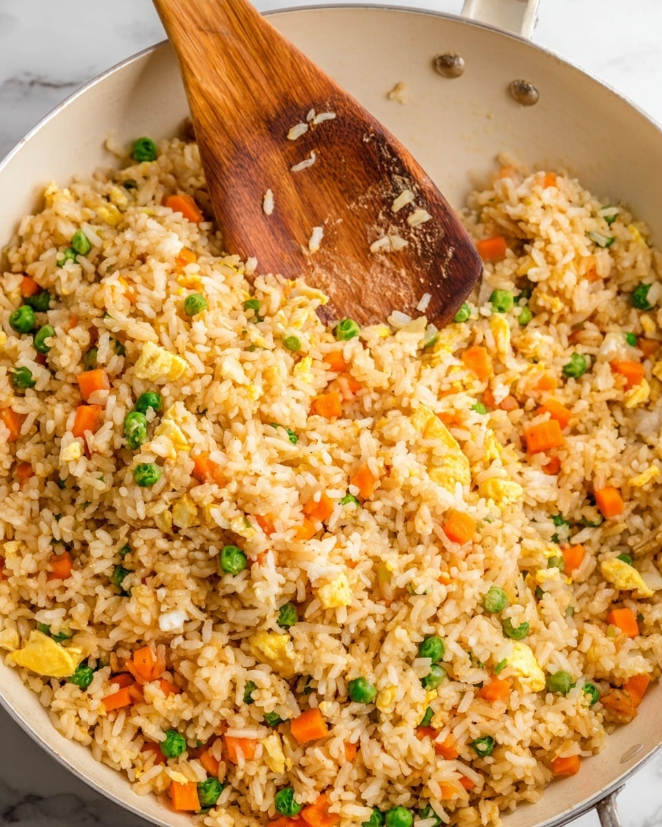 A close-up view of a large white pan filled with golden brown fried rice mixed with small bright orange carrot cubes and green peas scattered evenly throughout. The rice grains are separated and fluffy with bits of scrambled light yellow egg mixed in for extra texture. A wooden spatula rests on top, partially immersed in the rice, showing some grains stuck to its smooth surface. The pan sits on a white marbled textured surface, creating a clean and bright setting. Photo taken with an iphone --ar 4:5 --v 7