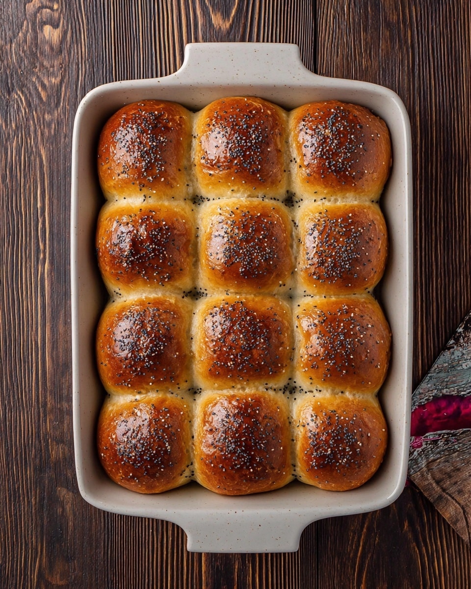 A white baking dish holds a set of 12 small, soft dinner rolls arranged in a 3 by 4 grid, each roll topped with a shiny golden-brown crust and sprinkled with black poppy seeds. The rolls are closely packed, showing slight browning at the edges where they touch. The dish is placed on a dark wooden surface with visible grain patterns. Part of a colorful food package is seen at the bottom right corner. photo taken with an iphone --ar 4:5 --v 7