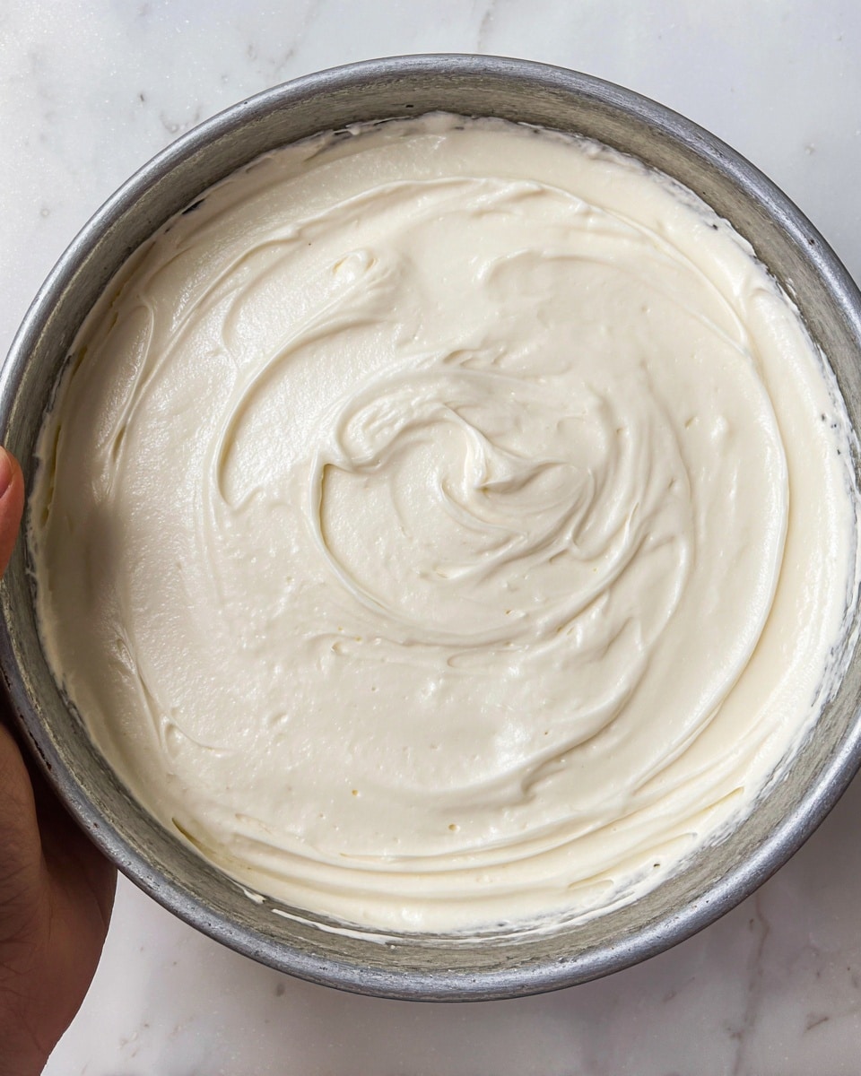 The image shows a single layer of smooth, creamy white frosting spread evenly inside a round metal cake pan. The frosting has a glossy texture with soft peaks and swirls, giving it a light and airy look. The cake pan sits on a white marbled surface, and a woman's hand holding the pan is partially visible on the left side. Photo taken with an iphone --ar 4:5 --v 7