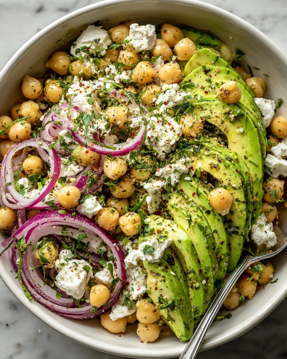 The image shows a white bowl filled with a fresh salad made of several layers. The bottom layer has bright green avocado slices with smooth and creamy texture, arranged in a slightly overlapping way. On top of the avocado, there are light beige chickpeas that look round and firm, scattered evenly everywhere. Thin rings of red onion with a glossy, translucent purple color are spread among the chickpeas and avocado. Crumbled white cheese pieces with a soft and crumbly texture are sprinkled across the salad. The dish is garnished with fresh green chopped herbs and a mixture of black and red seasoning, adding specks of color all over. A metal spoon is placed inside the bowl, partially visible on the right side. The bowl sits on a white marbled surface. Photo taken with an iphone --ar 4:5 --v 7
