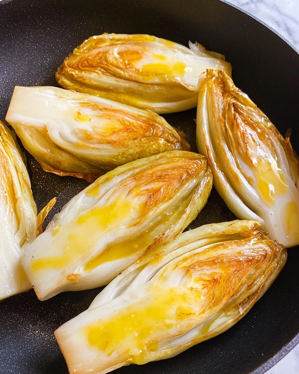 The image shows six pieces of cooked endive halves placed inside a black pan. Each piece has a glossy, slightly browned surface with shiny yellow spots from the cooking oil. The texture is soft and smooth, and the edges have a light crispness with slight browning. The endive pieces are arranged randomly but close together, covering most of the pan's bottom. The background beneath the pan is a white marbled texture photo taken with an iphone --ar 4:5 --v 7