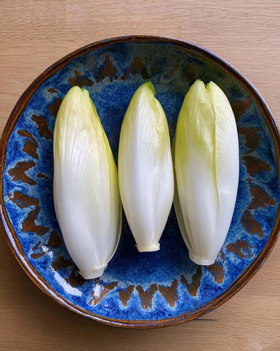 Three fresh small endive leaves are placed side by side on a white ceramic plate with deep blue and brown patterns. Each leaf forms a smooth, pale yellow-white to light green elongated shape, tapering at the top and thicker at the base. The plate rests on a light wood surface. photo taken with an iphone --ar 4:5 --v 7