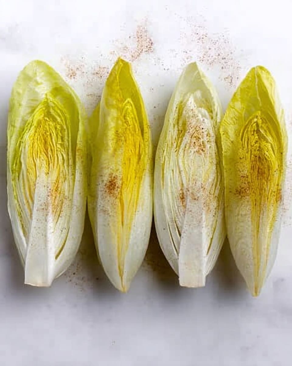 The image shows four halves of chicory leaves arranged in a row on a white marbled surface. Each leaf half has a smooth, shiny texture with a pale yellow-green color, showing some veins and natural curves. Two of the halves show more yellow-green hues in the center, while the other two are mostly white with a slight green tint near the edges. A light brown powder is sprinkled lightly over all the leaves. The clean, simple setting highlights the fresh and natural look of the chicory leaves photo taken with an iphone --ar 4:5 --v 7