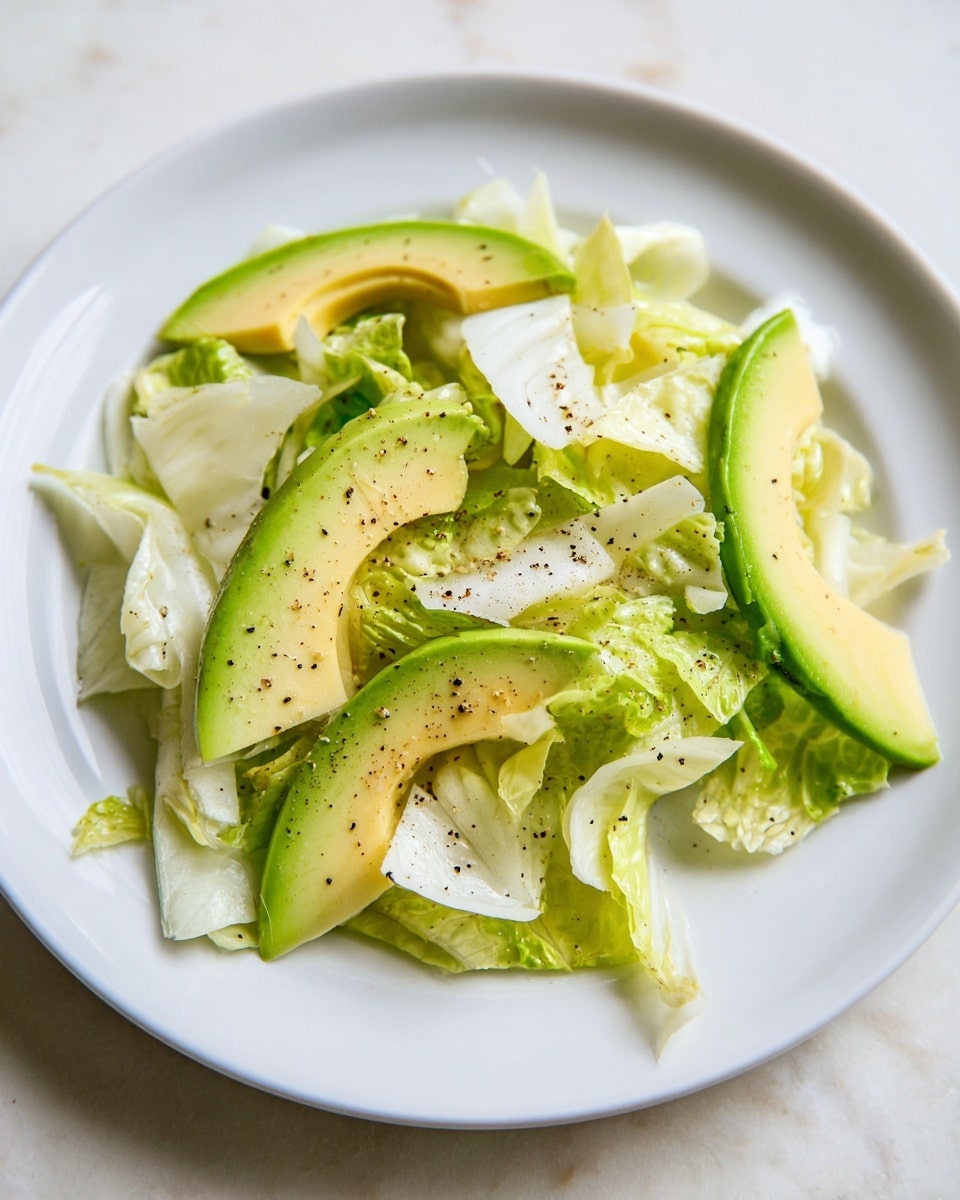 A simple salad is placed on a white plate, resting on a white marbled surface. The salad has two main layers: the first layer is made of pale green and white lettuce pieces with soft, curly textures scattered across the plate. On top of this lettuce base, there are several slices of ripe avocado showing smooth, creamy, light green flesh with darker green edges. Black pepper specks are scattered lightly over the salad, adding small dark spots on the light greens and whites of the ingredients. The overall look is fresh and light with natural green and white colors. photo taken with an iphone --ar 4:5 --v 7