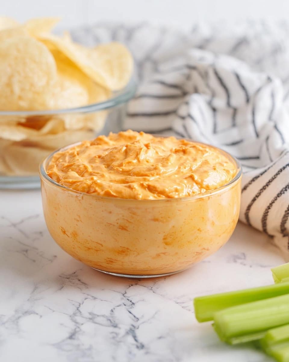 A clear glass bowl filled with a thick, creamy orange dip that has a slightly rough texture and small visible chunks, sitting on a white marbled surface. Behind the bowl, on the left, there is a clear glass bowl filled with light beige potato chips. On the right side, there is a soft, white fabric with black stripes, slightly crumpled. In the front right corner, bright green celery sticks rest partially in the frame. photo taken with an iphone --ar 4:5 --v 7