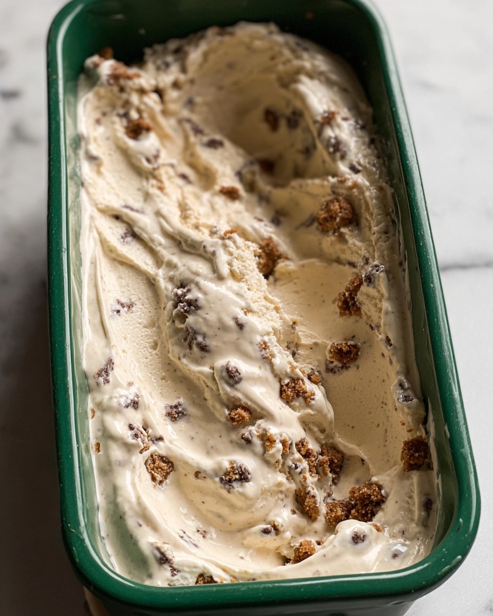 A close-up view of a single thick layer of light beige ice cream with small dark specks and crushed brown cookie pieces scattered unevenly on top and mixed throughout. The ice cream is inside a rectangular green container, with smoothed and slightly swirled texture, some ice cream slightly spread up the sides. The container sits on a white marbled surface. Photo taken with an iphone --ar 4:5 --v 7